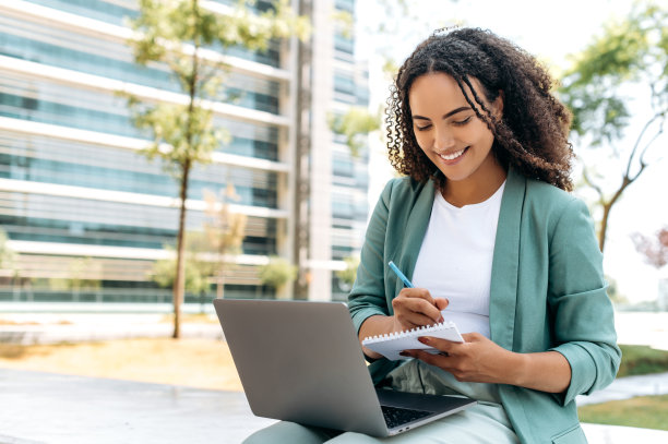 在线研讨会。聪明成功的巴西或拉丁裔年轻女性，大学生，自由职业者，坐在户外的笔记本电脑，听在线讲座，在笔记本上写下信息，微笑图片下载