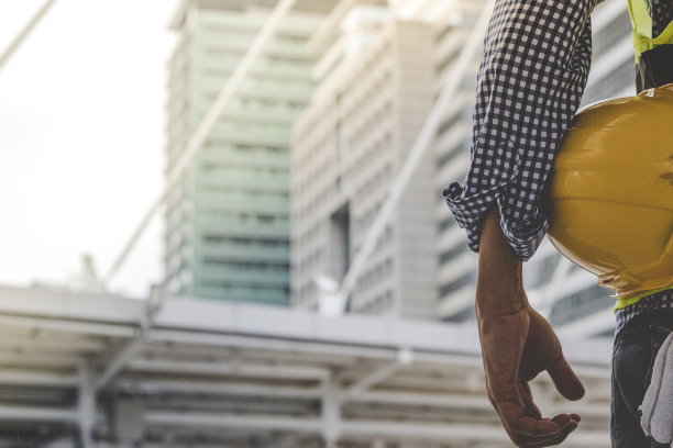 工程师手拿安全帽白色工作头盔，为土木建筑工程安全帽。在工地上，身穿安全服的建筑工程师手持白色工作头盔和安全帽图片下载