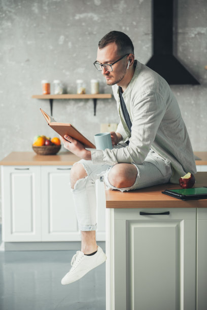 商人在厨房里看书喝茶。使用电子设备的闲暇时间。在室内享受早餐咖啡。图片下载