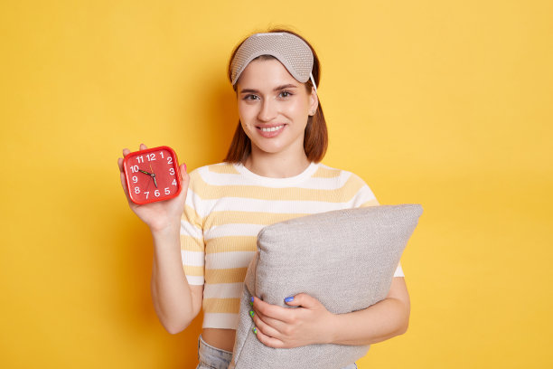 年轻的微笑快乐的女人穿着t恤和眼罩，放松在家里拿着闹钟隔离在黄色背景，看着相机与积极的面部表情。图片下载