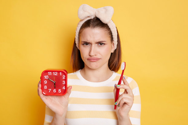 室内悲伤不安的女人穿着休闲风格的条纹t恤，白色的牙刷和红色的闹钟，摆出孤立的黄色背景。口腔卫生。图片下载
