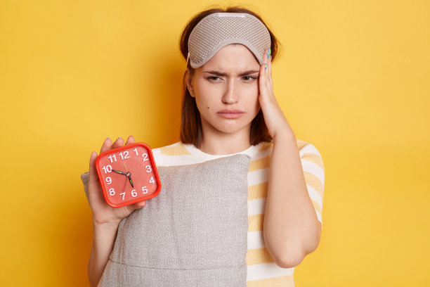 室内拍摄的疲惫不健康的女性，穿着条纹衬衫，戴着睡眠面罩，抱着枕头和红色闹钟，睡眠不足，头部疼痛，在黄色背景下孤立地摆姿势。图片下载