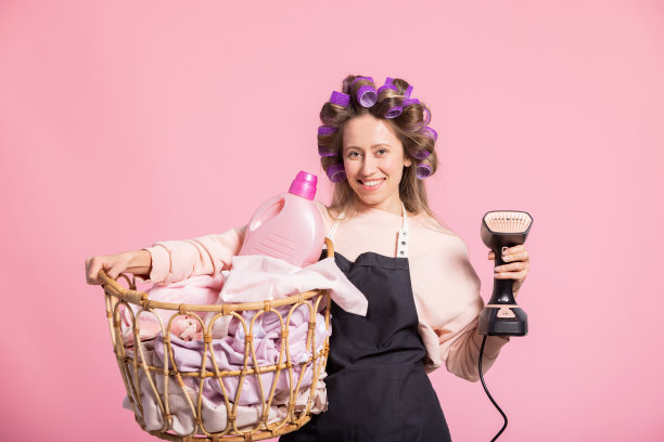 一个快乐的女人打扫人们的家作为一个清洁工工作。家佣插入洗衣，熨烫衣服的围裙与辊在头部肖像的粉红色背景图片下载