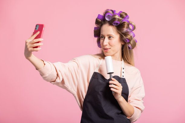 粉色背景上的女人肖像。头戴卷发卷的女孩微笑着对着洗衣服卷唱歌录视频，对着相机打电话图片下载