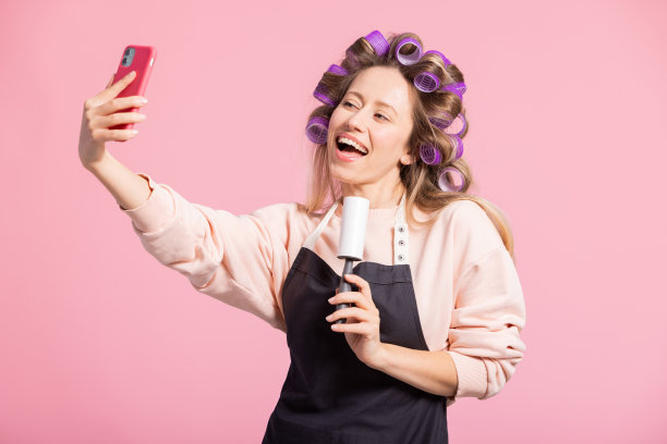 粉色背景上的女人肖像。头戴卷发卷的女孩微笑着对着洗衣服卷唱歌录视频，对着相机打电话图片下载