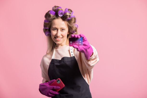 面带微笑的女子与卷发在头上的姿势在粉红色的背景下，手里拿着橡胶手套保护的信用卡，订购清洁配件，在手机上的应用程序支付图片下载