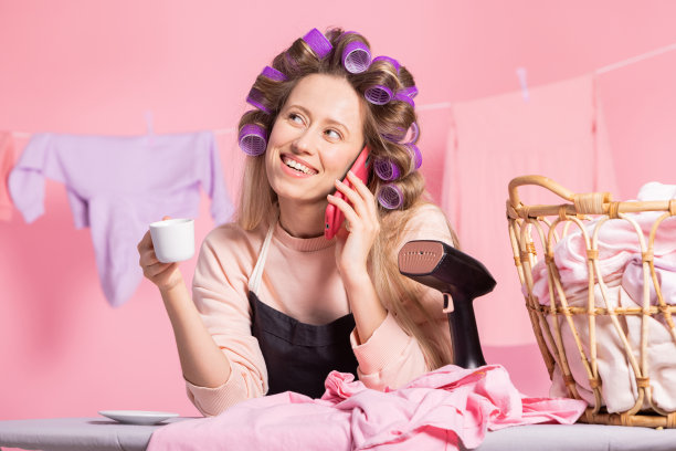 照片上的女人一边喝着咖啡一边打电话，背景是粉红色的衣服。做家务，熨衣服，挂衣服准备出门图片下载