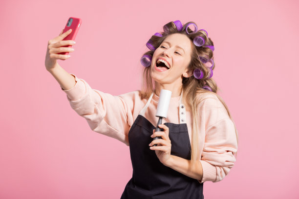 这位快乐的家庭主妇整理公寓，对着用卷发器涂在头发上的衣服滚筒唱歌，自拍，并在社交媒体上录制视频。肖像微笑的女人在粉红色的背景图片下载