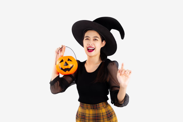 万圣节的主题，年轻漂亮的亚洲妇女在黑色万圣节服装戴女巫帽拿着橙色的南瓜桶摆指着白色的背景图片下载