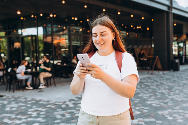 年轻女子在户外的街道上用手机聊天的形象。背着背包的年轻女性在城市里使用智能手机。城市生活方式的概念。图片下载