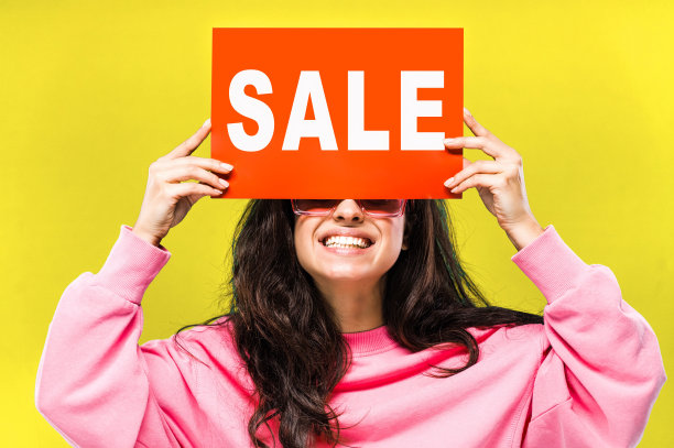 大的销售。迷人的白人年轻长头发妇女手持红色的“出售”标志在她的头附近和微笑，在黄色孤立的背景图片下载