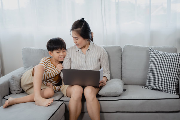 英俊的亚洲男孩跑到他的妈妈身边坐下，而他的妈妈正在用笔记本电脑。他的妈妈转过身来深情地看着她的儿子，开心地笑了。图片下载