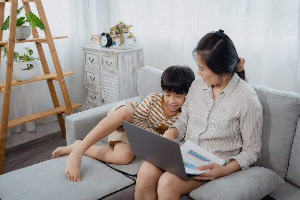 英俊的亚洲男孩跑到他的妈妈身边坐下，而他的妈妈正在用笔记本电脑。他的妈妈深情地看着他，轻轻地抚摸着儿子的头。图片下载