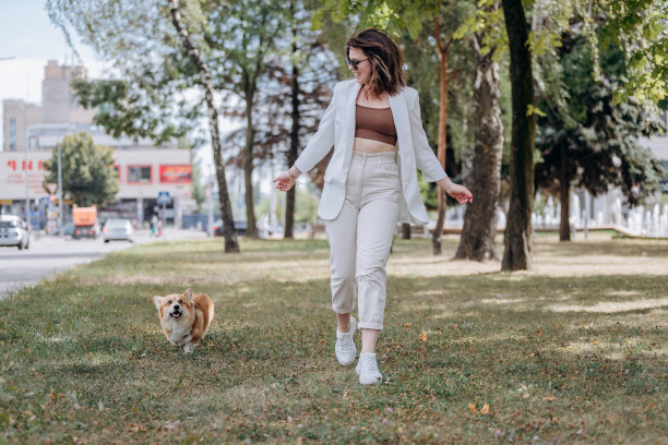 快乐的女人穿着白色西装，戴着太阳镜，和威尔士柯基犬在城市公园散步图片下载