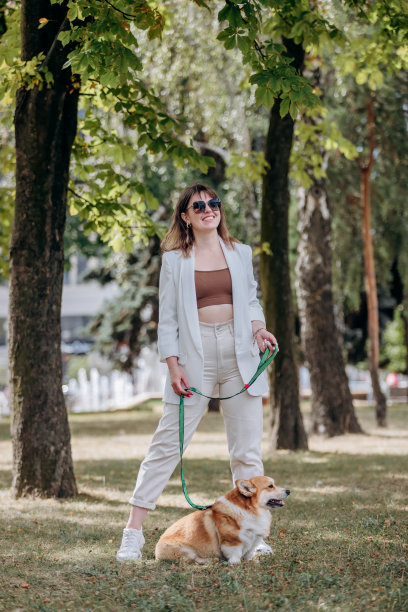 快乐的女人穿着白色西装，戴着太阳镜，和威尔士柯基犬在城市公园散步图片下载