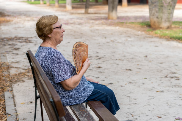 侧视图。白人老年妇女坐在公园长凳上扇着扇子图片下载
