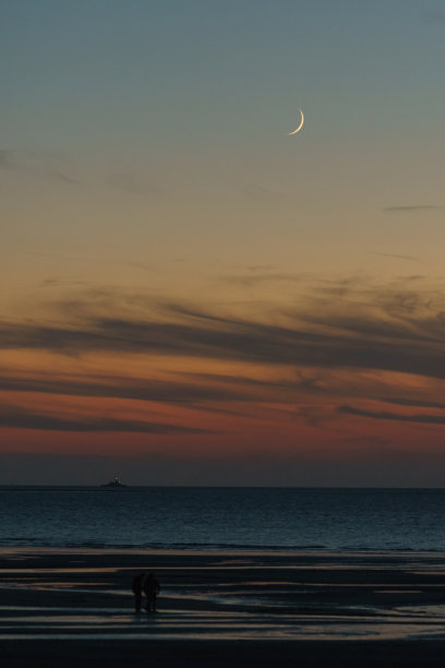 黄昏时海上的新月，远处的船和日落后海滩上行人的剪影图片下载