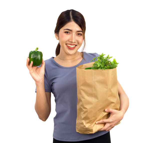 穿着便装的年轻亚洲女子微笑着站着，手里拿着青椒，另一只手拿着装满蔬菜的纸袋。肖像在白色背景与工作室的光。孤立的图片下载
