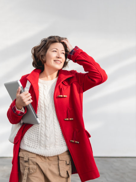 穿着红外套的卷毛女人站在白墙旁边。带着笔记本电脑微笑的成年学生。自由职业者与便携式设备的工作。现代的生活方式。图片下载
