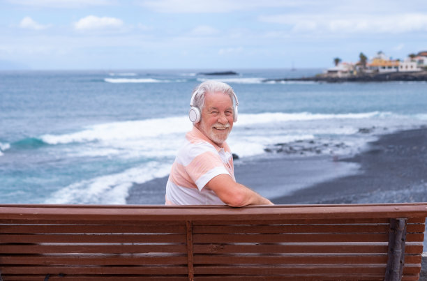欢快的大胡子老人坐在海边的长凳上，戴着耳机听着音乐，老人享受着自由和退休，地平线上的水图片下载