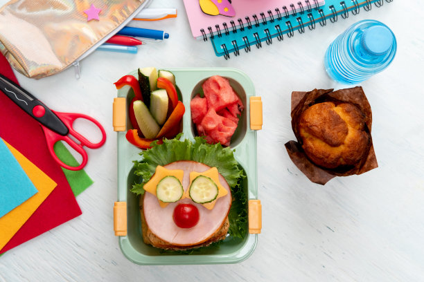 学校为孩子们准备的午餐盒，食物是滑稽的面孔。学校午餐盒，桌上有三明治、蔬菜、水和文具。健康的饮食习惯概念。回到学校的概念图片下载