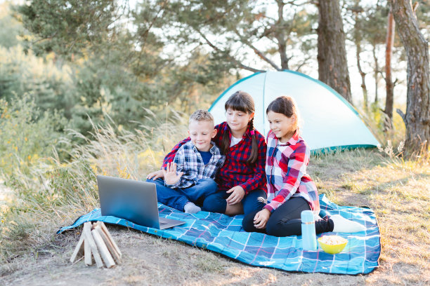 孩子们在森林里看笔记本电脑。男孩和女孩正在帐篷营里休息。夏天，孩子们坐在草地上的格子上。图片下载