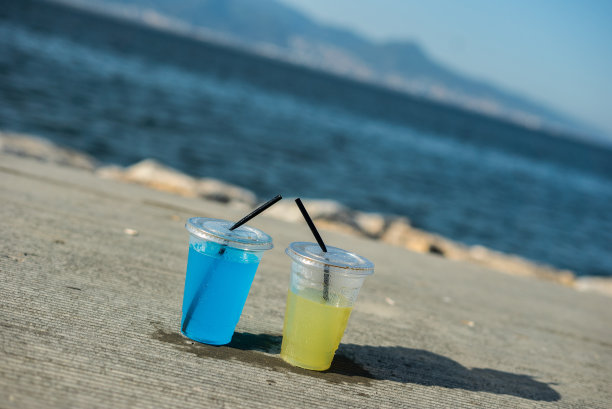 透明塑料杯是黄色和蓝色的饮料是在桌子上的户外焦点对前景垂直照片海是背景图片下载