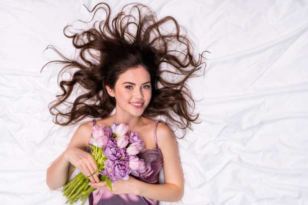 上面高角视角肖像迷人的时髦快乐的女孩躺在白色舒适的亚麻布拿着鲜花植物亲密图片下载