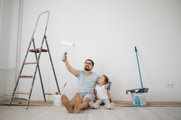 年轻的父亲和他的金发可爱的小女孩女儿与墙壁油漆辊坐在地板上图片下载