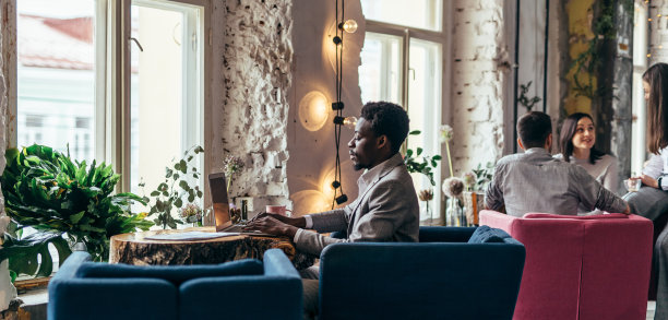 一名非裔美国男子坐在咖啡桌旁使用笔记本电脑，在远处工作或学习。图片下载