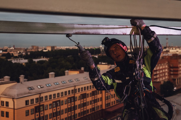 工业夜城工作理念。工业登山工人穿着制服，在黄昏的城市背景下，悬挂在建筑物和油漆辊施工上，油漆立面。复制文本空间图片下载