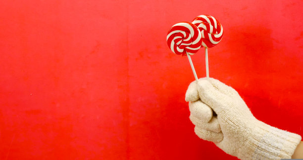 他们一只手戴着冬天的手套，背景是红色，手里拿着两个薄荷和焦糖味的圆形棒棒糖。有一个地方可以插入贺词，圣诞背景和明信片。图片下载