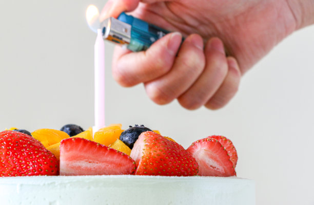 水果生日蛋糕的特写，上面有新鲜的草莓，芒果和蓝莓。在背景的焦点之外，一只手轻弹打火机，点燃有火焰的粉红色蜡烛图片下载
