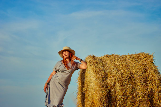 一个年轻漂亮的女人，戴着帽子，穿着夏装，正坐在田野里的一捆干草上。乡村自然，麦田，休养于乡，与自然合一图片下载