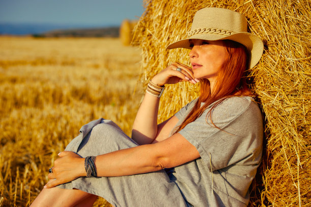 一个年轻漂亮的女人，戴着帽子，穿着夏装，正坐在田野里的一捆干草上。乡村自然，麦田，休养于乡，与自然合一图片下载