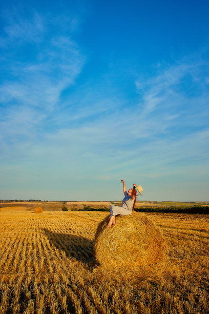 一个年轻漂亮的女人，戴着帽子，穿着夏装，正坐在田野里的一捆干草上。乡村自然，麦田，休养于乡，与自然合一图片下载