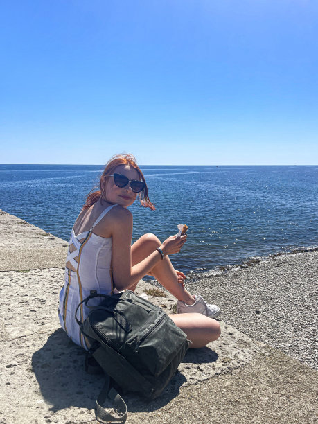 特写形象的女人红头发穿太阳裙，镜子太阳镜与背包，坐在散步在有风的日子，吃冰淇淋，看着相机，蓝色的夏天的天空，港口地区在科布，莱姆里吉斯，多塞特，英国图片下载