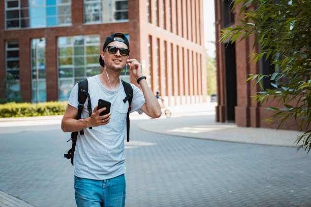 年轻的男人是一个时髦的时髦的人，戴着墨镜和黑色的帽子走在城市里，手里拿着智能手机，听着音乐耳机。T图片下载