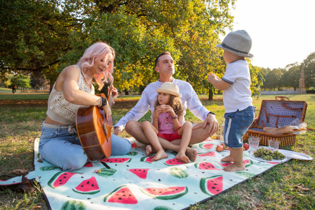 一个快乐的素食家庭在野餐，妈妈弹吉他，爸爸和孩子们玩，水果和素食食物放在野餐毯子上图片下载