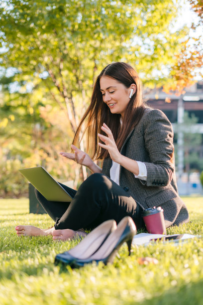 年轻的白人女商人正在笔记本电脑上进行在线视频通话，坐在户外的城市公园的绿色草坪上，微笑和手势。互联网通信，无线连接技术。业务电话。图片下载