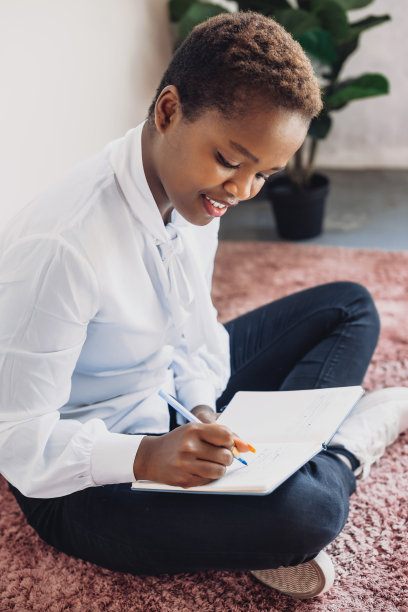 漂亮的非洲女人坐在地板上写笔记本，独自在家学习。现代商业的概念。人们生活方式的概念。用铅笔做记号，准备期末考试图片下载