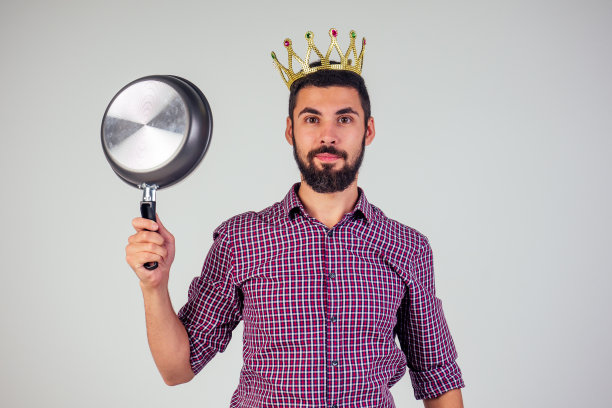 在画室的白色背景下，身穿格纹红衬衫、头戴金冠的英俊男子手拿金属煎锅。厨房之王，有创意又有趣图片下载