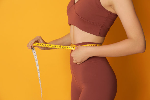 身着运动服的女子用胶带在黄色背景上测量腰部，特写图片下载