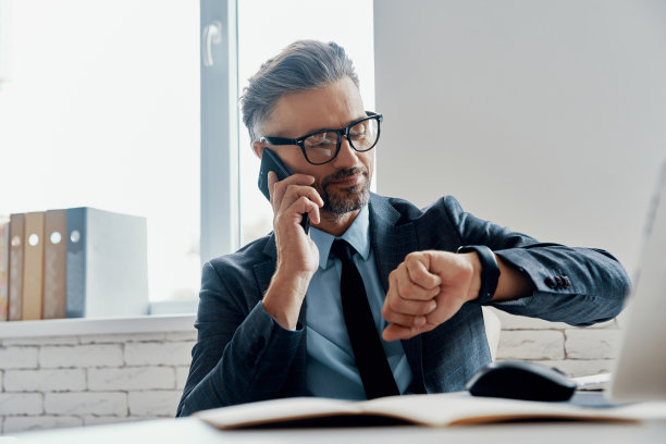 穿着正装的英俊成熟男人一边看时间一边在办公室打电话图片下载