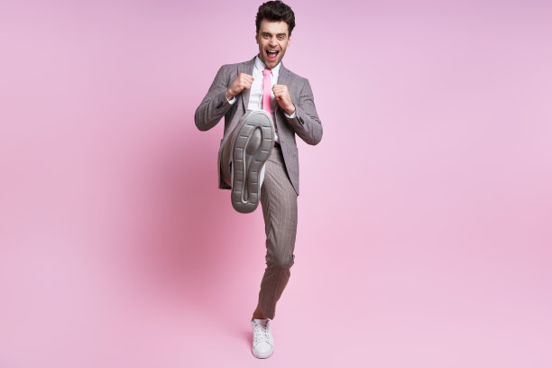 愤怒的年轻人在全套西装扔腿踢，站在粉红色的背景图片下载