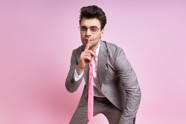 自信的男人穿着全套西装，把手指放在嘴唇上，站在粉红色的背景下图片下载