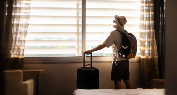 男游客戴着帽子，背着背包站在酒店房间里，手里拿着旅行袋。图片下载