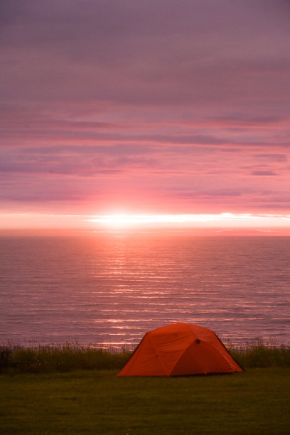 在海边的草地上搭起探险帐篷，欣赏壮观的粉红色午夜夕阳图片下载
