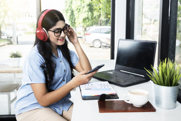 亚洲妇女戴着眼镜，用手机和耳机，笔记本电脑和笔记本电脑，桌上有一杯咖啡图片下载