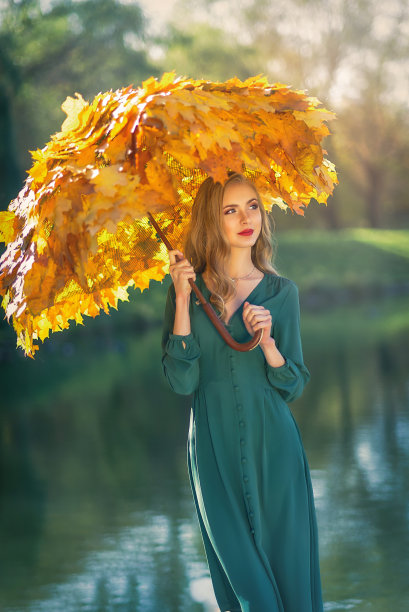 在一个秋天的公园里，一个年轻美丽的女人站在一把树叶做的伞下，背景是一个湖。美丽的女子在秋天的公园里散步，撑着枫叶的伞。图片下载